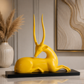 A glossy yellow deer sculpture lying down on a black rectangular base, displayed on a marble console table in an elegant interior setting with a vase of pampas grass and abstract wall art.