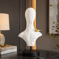 A white ribbed decorative bust sculpture with a small gold bird accent, displayed on a black stand on a dark marble table in an interior setting.